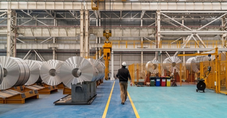 A man walks among large aluminum rolls in a factory.